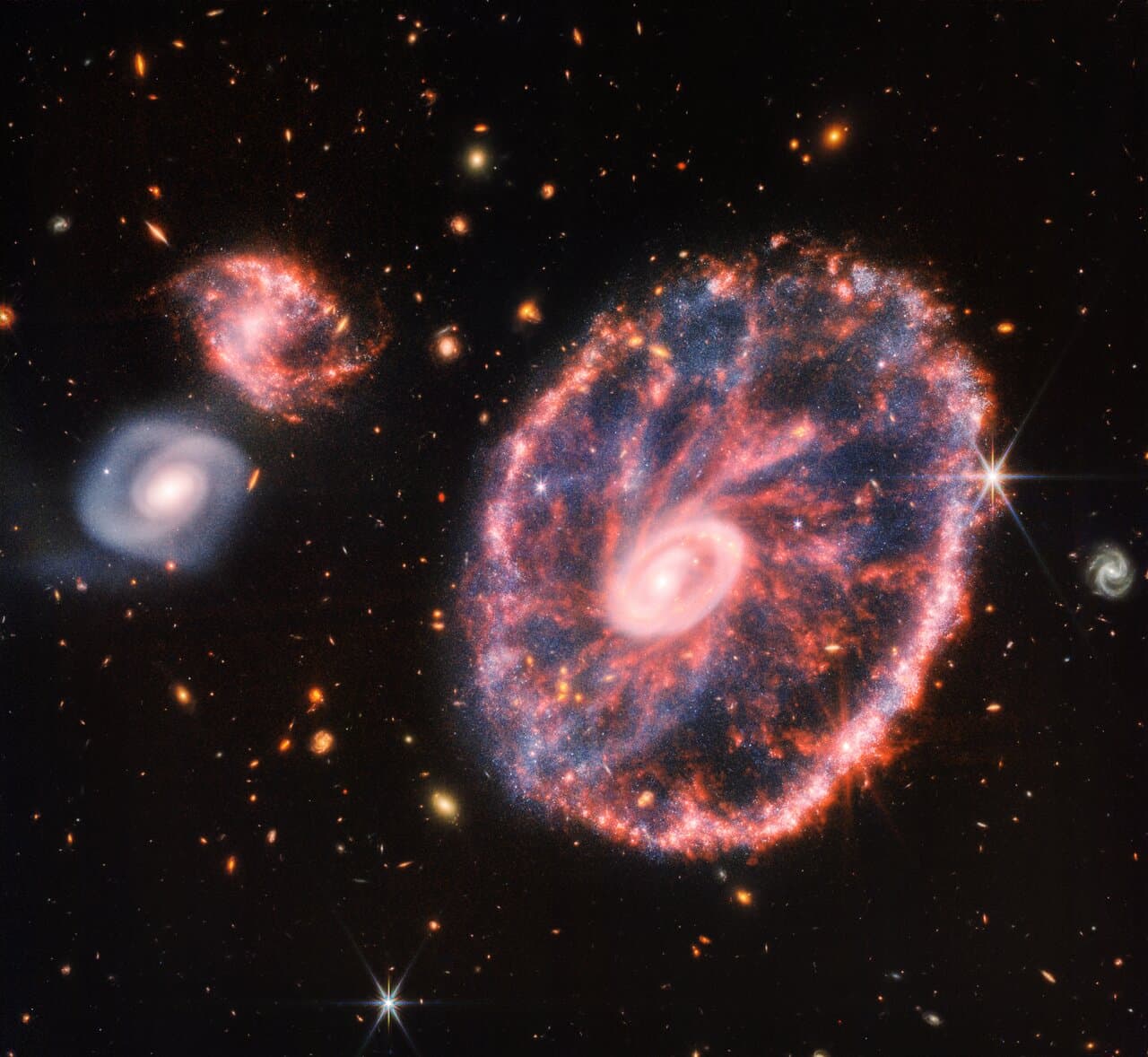 A galaxy shaped like a wagon wheel, with a bright inner ring, a colorful outer ring and red spokes between them, with a smaller spiral galaxy at the top left