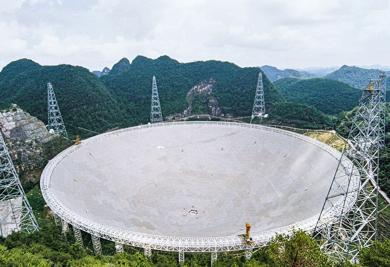 A huge round dish telescope set into a bowl of green hills, with cables and towers holding a receiver above it