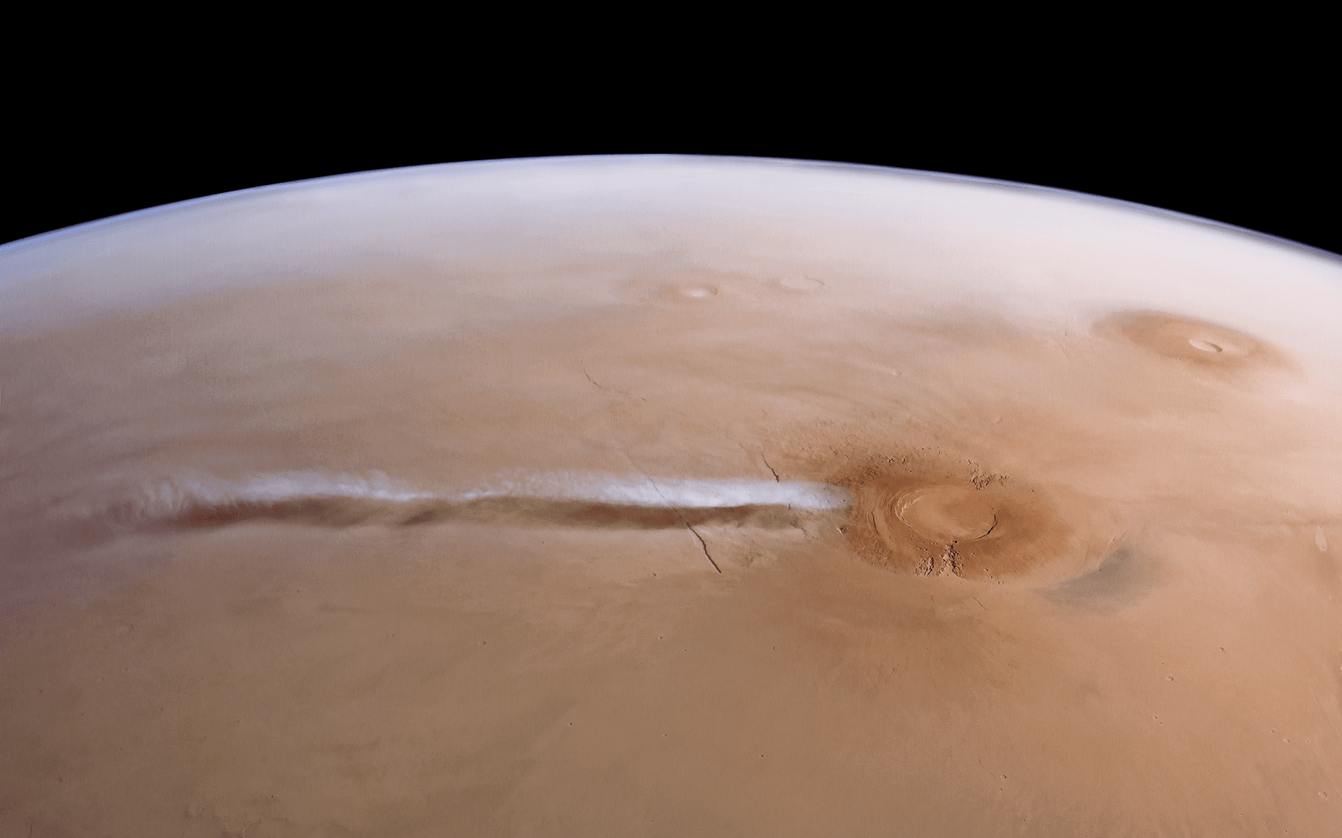 A tan Martian landscape seen at an angle from orbit, with a volcano at lower right and a long, thin white cloud trailing from it to the left, its shadow visible on the ground below